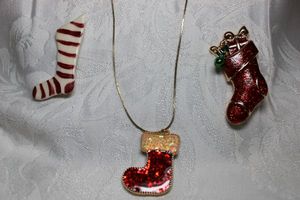 lot 268 image: (3) Holiday stocking jewelry. Includes an enamel broochpin 2.25H, a kiln fired clay hand painted stocking pin 2.25H and a glittered enamel stocking pendant on 24L adjustable length necklace.