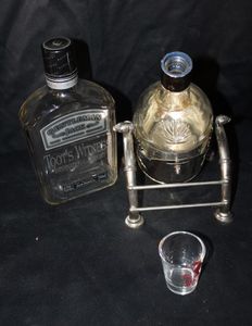 lot 154 image: Vintage Jack Daniels decanter set featuring two distinct empty bottles- a gentlemen Jack style bottle and a rounded cameo glass bottle paired with a metal stand and shot glass