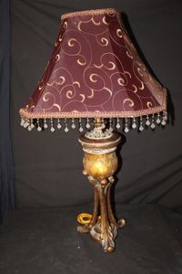 lot 331 image: Ornate Table lamp featuring a bell-shaped cranberry or burgundy-colored fabric shade adorned with a scrollwork pattern with a beaded fringe. Amber Colored Glass and faux wood or metal bronzed finished tall base,&nbsp