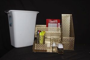 lot 128 image: Desk Set up in gold, paper holdersorganizers, white stapler, box of staples, new pens, and highlighters and various pens, White trashcan and gold tape dispenser