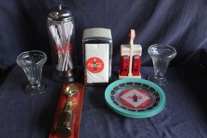 lot 8 image: Vintage Coke table items, Salt and pepper shaker, napkin holder and napkins, ice cream scoop in package, plates and straw holder, two clear glass sundae dishes