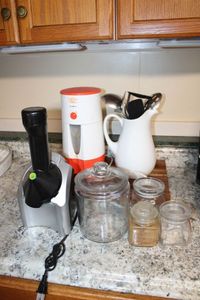 lot 34 image: Clear Glass Cannisters, Wood Cutting Board, Healthy Foods Juicer Model 901, Mr. Coffee Iced Tea Maker, and Pitcher with Utensils